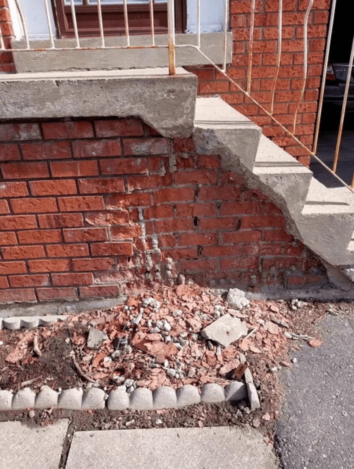 Spalling red brick and deteriorated mortar beneath concrete steps, with loose masonry debris along the foundation wall