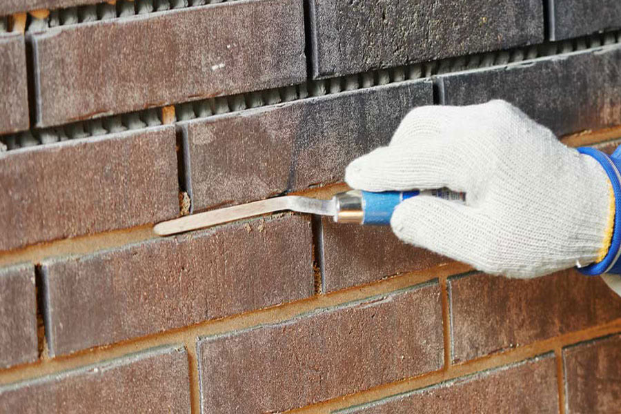 Gloved mason uses a jointing tool to work mortar between brown brick courses