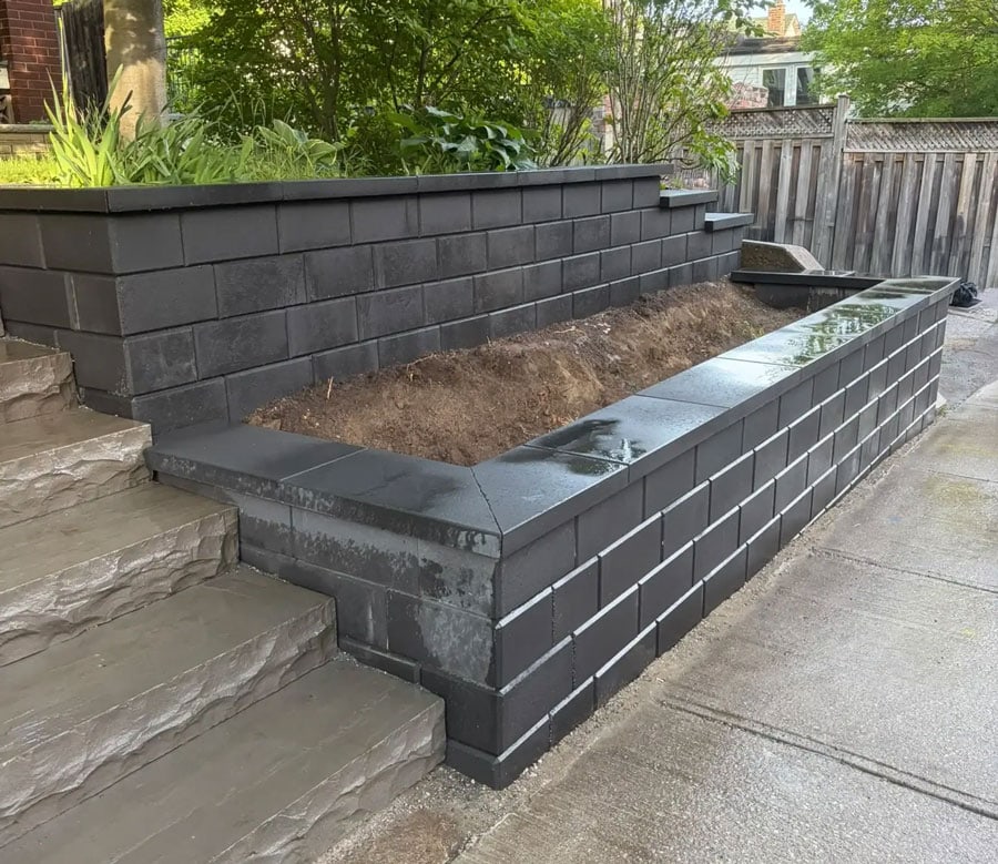 Black block retaining wall with capped edges beside stone steps and a concrete walkway