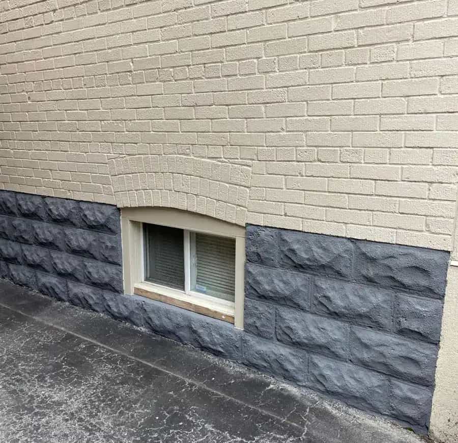 Dark gray coated foundation wall beneath a light-painted brick wall and basement window