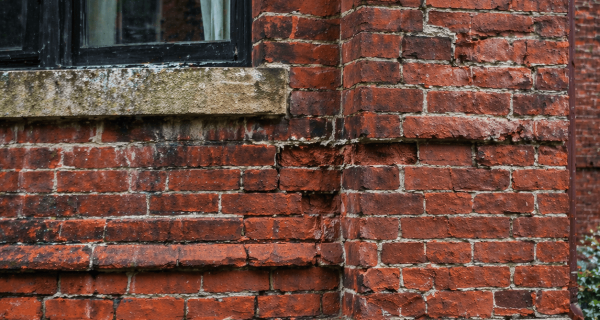 Weathered red brick wall with a black-framed window, stone sills, recessed mortar, and visibly deteriorated bricks