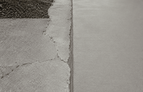 Cracked concrete walkway beside a smoother concrete path near a stone-faced wall and downspout
