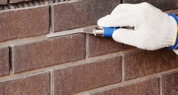 Gloved mason uses a pointing trowel along a mortar joint in a brick wall