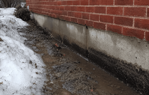 Muddy meltwater pooling beside a brick wall and concrete foundation during winter thaw