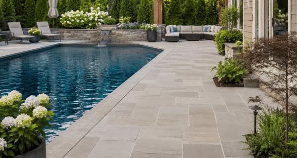 Light stone patio surrounding a backyard swimming pool with landscaped planting beds and a pergola
