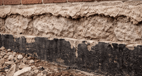Weathered brick wall above a rough exposed foundation with peeling coating and loose stones along the ground