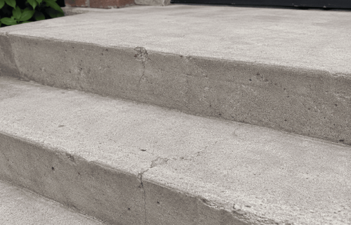 Concrete entrance steps with visible cracks, chipped edges, and a brick wall beside a black door