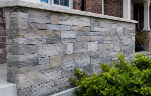 Gray stone veneer wall with a light masonry cap beside a residential entrance and landscaped planting bed