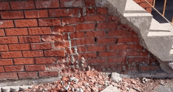 Spalling red brick and deteriorated mortar beneath concrete steps, with loose masonry debris along the foundation wall